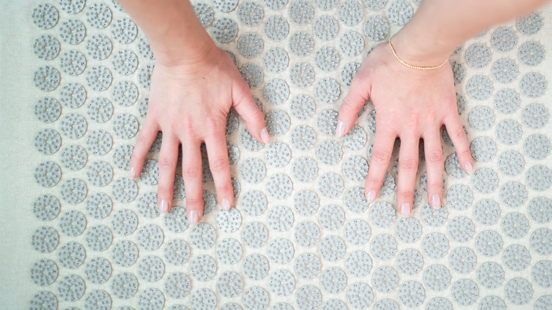 Nahaufnahme von zwei Händen, die auf einer Akupressurmatte liegen. Die Matte ist mit runden, stacheligen Massagepunkten ausgestattet, die zur Stimulation der Handflächen und Förderung der Durchblutung dienen. Die Person trägt ein schlichtes Armband und die Szene zeigt einen Moment der Entspannung oder Akupressurbehandlung.
