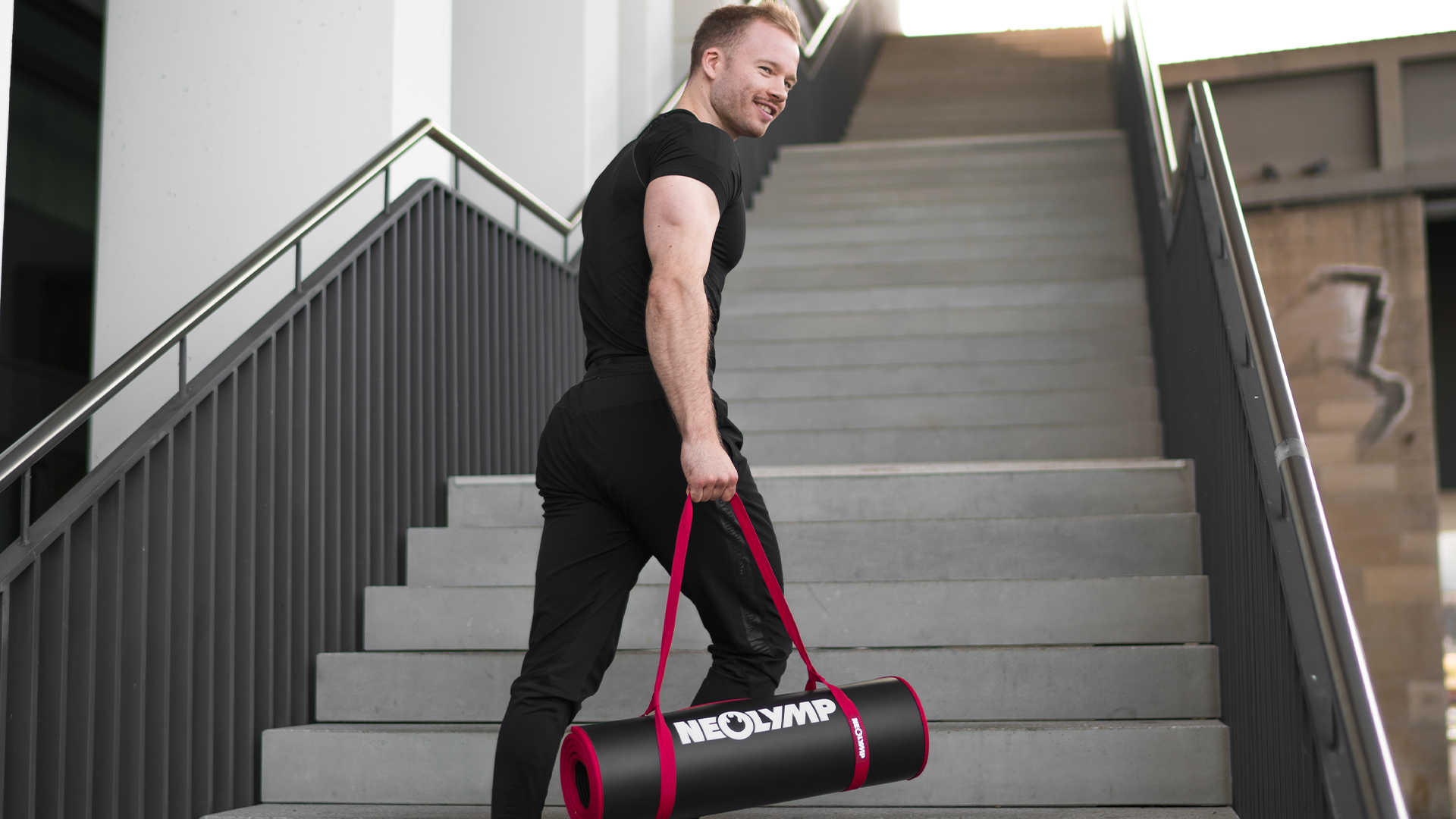 Mann in schwarzer Sportbekleidung trägt eine NEOLYMP Trainingsmatte auf einer Treppe. Perfekt für das Workout unterwegs.