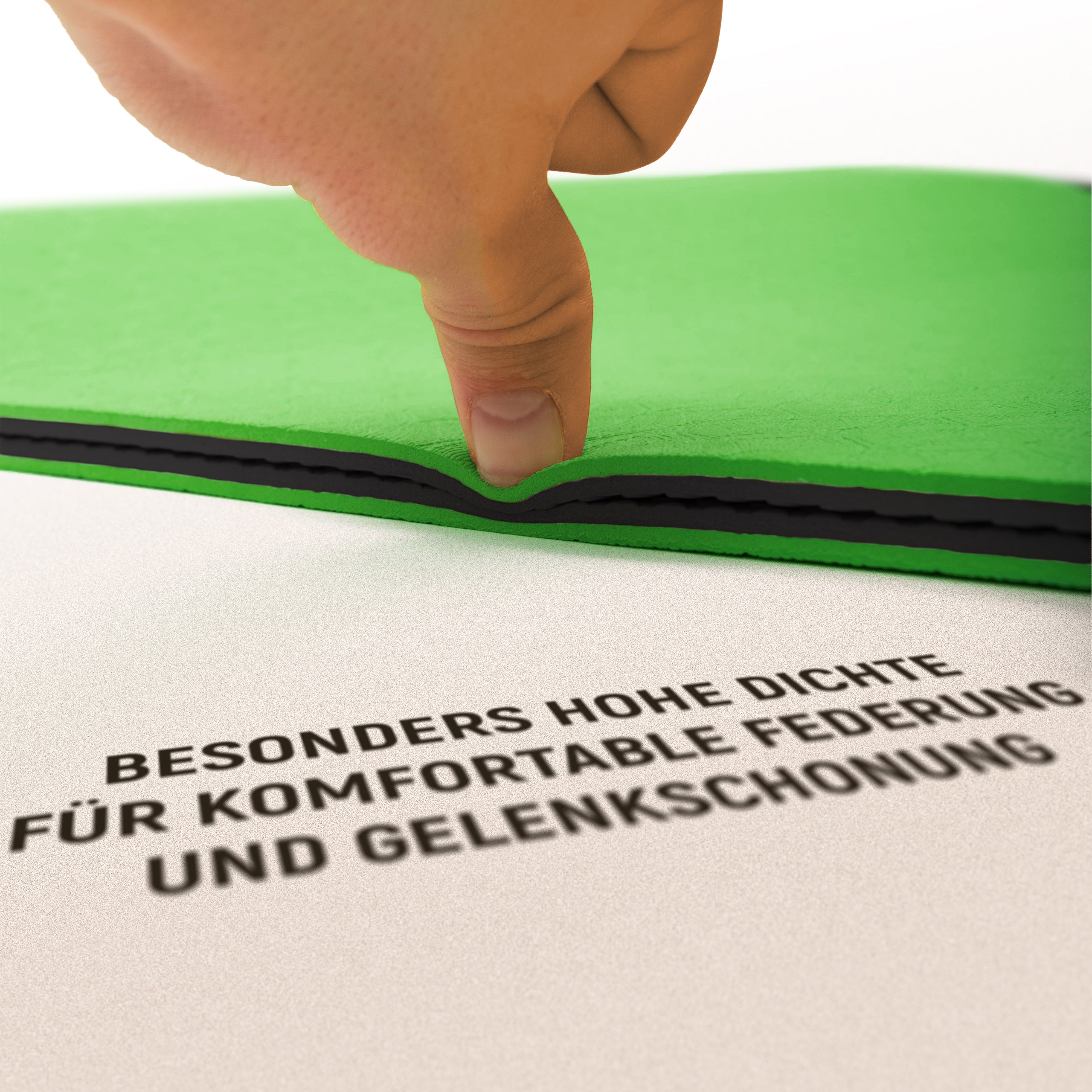 Eine Nahaufnahme einer grünen Trainingsmatte, die von einer Hand zusammengedrückt wird, um ihre hohe Dichte und Federung zu demonstrieren. Der Text auf der Matte lautet: "BESONDERS HOHE DICHTE FÜR KOMFORTABLE FEDERUNG UND GELENKSCHONUNG." Die Abbildung hebt die Qualität der Matte hervor, die für ein schonendes und komfortables Training sorgt.
