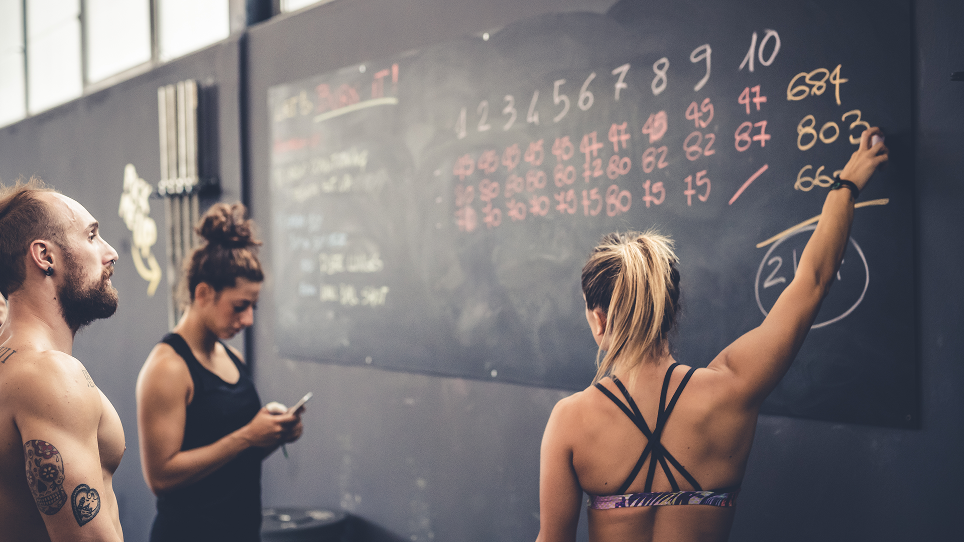 Drei Personen in einem Fitnessstudio stehen vor einer Tafel mit Trainingspunkten. Eine Frau schreibt Ergebnisse auf die Tafel, während eine andere Person auf ihr Handy schaut. Ein Mann mit tätowiertem Oberarm beobachtet das Geschehen aufmerksam. Die Tafel zeigt verschiedene Zahlen und Notizen zu den Trainingseinheiten an.