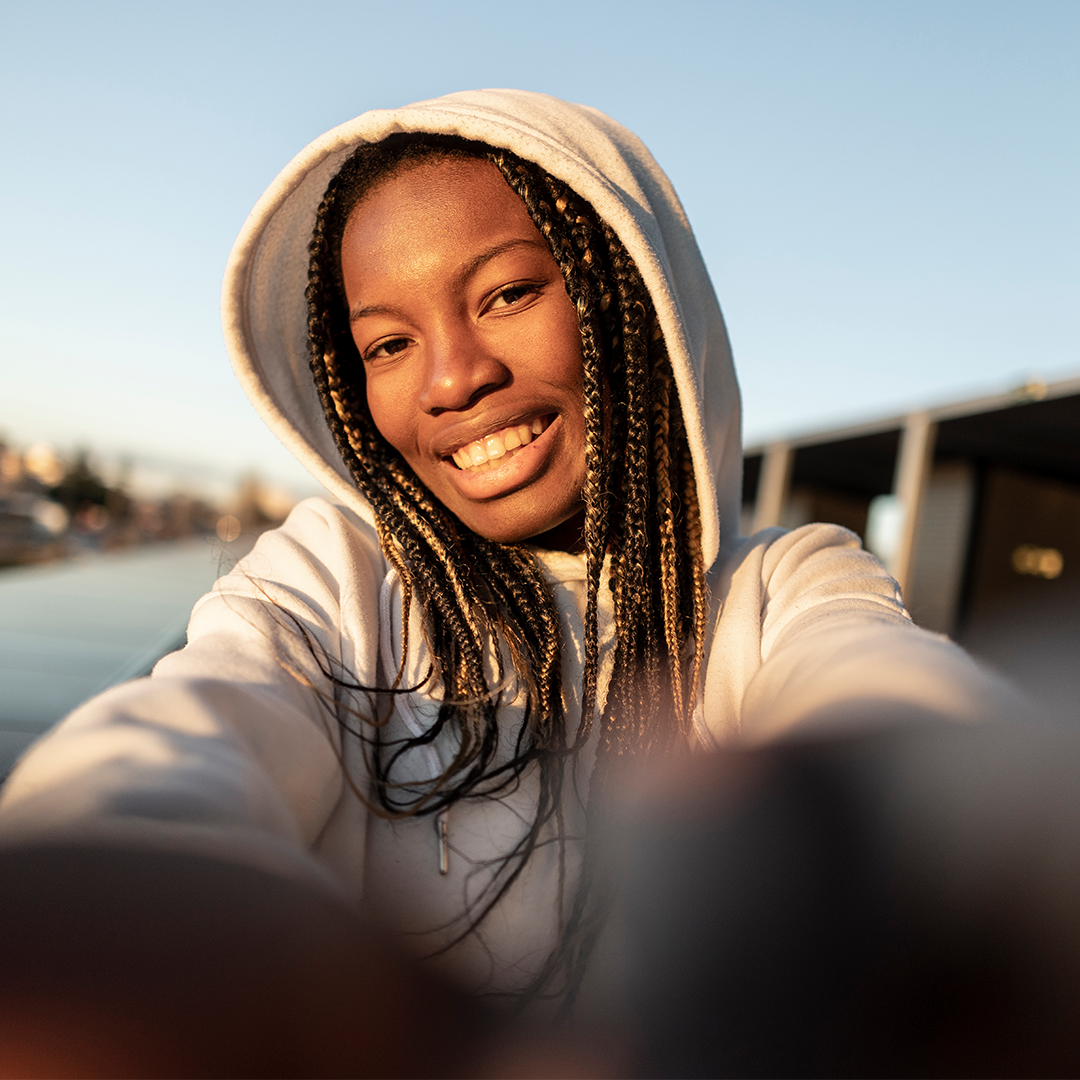 Eine junge Frau mit geflochtenen Haaren trägt einen hellen Hoodie und lächelt in die Kamera, während sie ein Selfie macht. Die Sonne geht unter und beleuchtet ihr Gesicht warm, was eine entspannte und fröhliche Atmosphäre schafft. Im Hintergrund sind unscharfe Gebäude und ein klarer Himmel zu sehen.