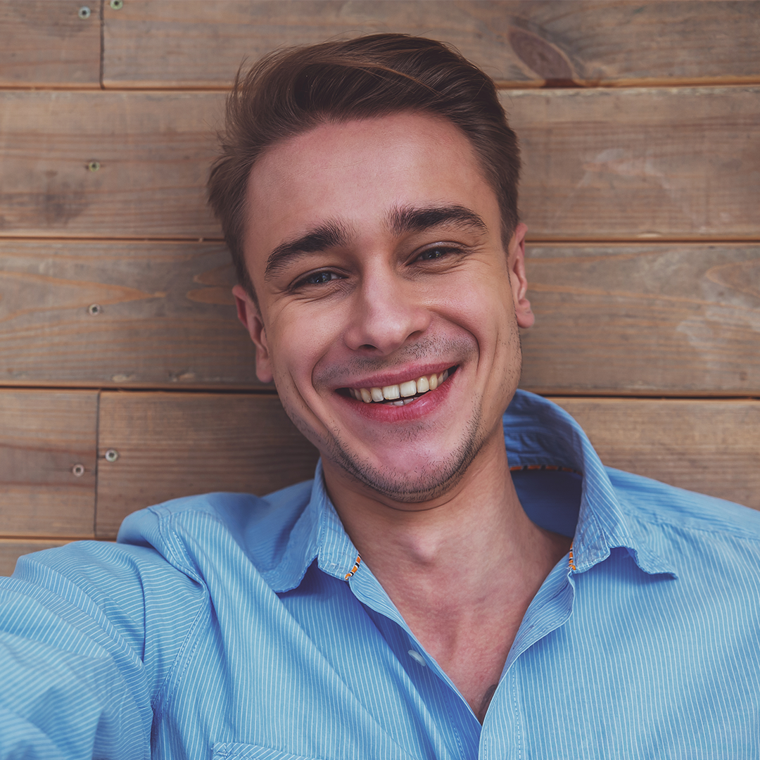 Ein junger Mann in einem hellblauen Hemd lächelt herzlich in die Kamera. Er liegt auf einem hölzernen Hintergrund und wirkt entspannt und glücklich.