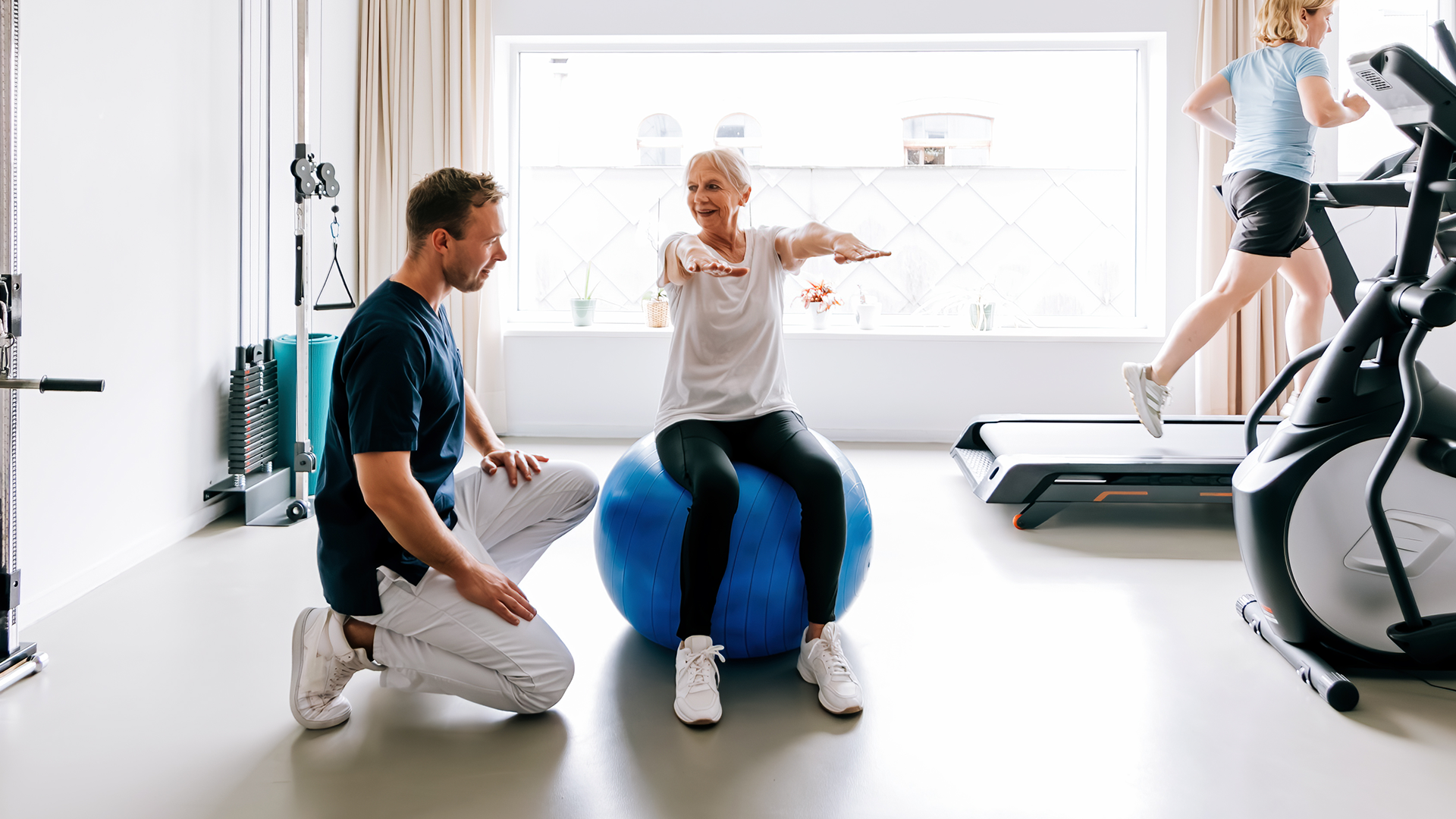 Personal Trainer unterstützt ältere Frau bei einem Balance- und Krafttraining auf einem blauen Gymnastikball, während eine weitere Person im Hintergrund auf einem Laufband trainiert.