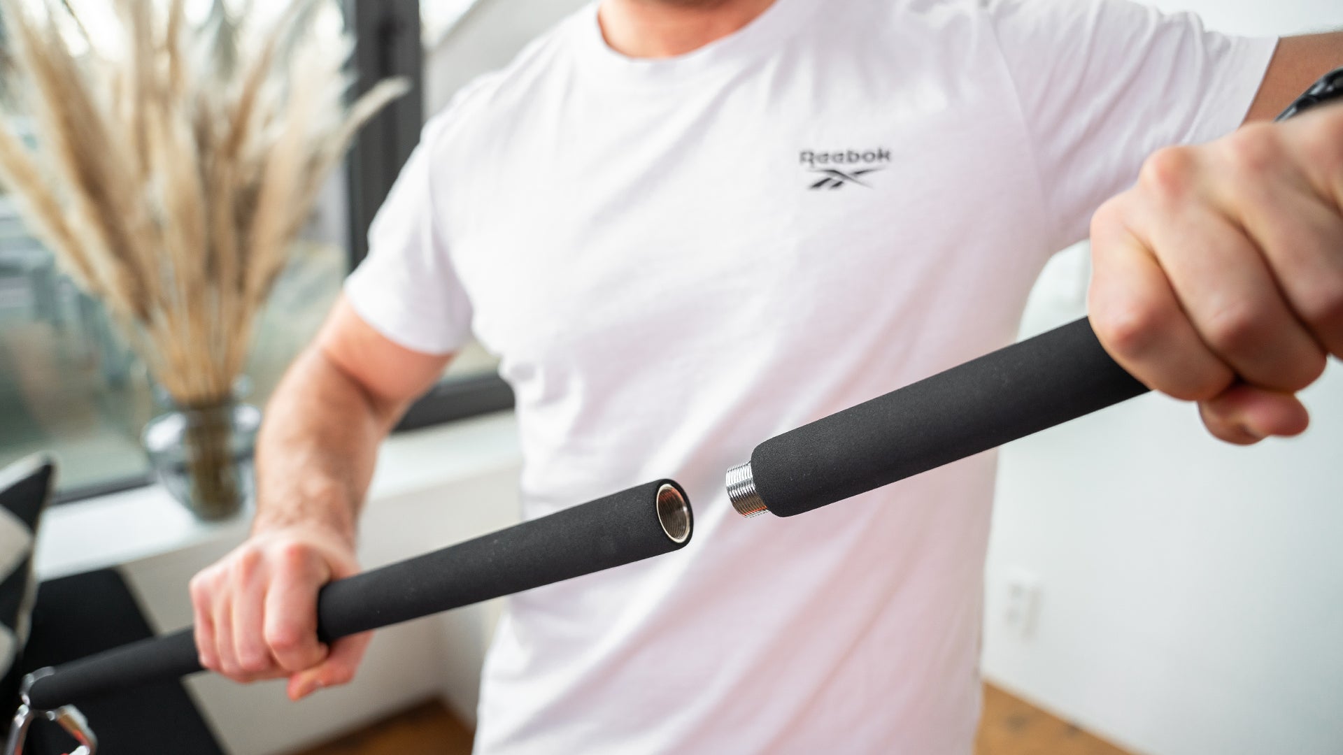 Ein Mann in einem weißen Reebok-T-Shirt hält zwei Enden einer Stange, die er auseinanderzieht oder zusammenschraubt. Die Stange hat eine schwarze, gepolsterte Oberfläche und Metallverbindungen. Im Hintergrund ist eine moderne Inneneinrichtung mit einer Vase und dekorativen Pflanzen zu sehen.