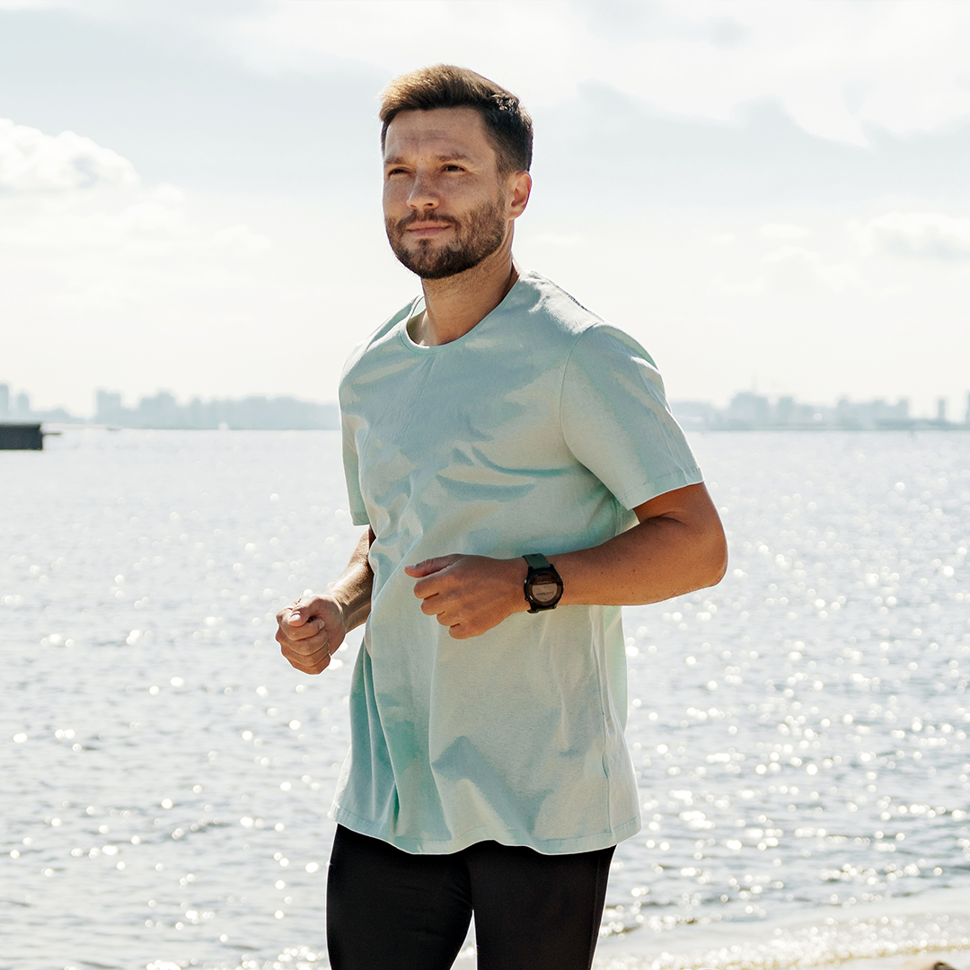 Ein Mann joggt am Ufer eines Gewässers entlang, gekleidet in ein helles T-Shirt und eine dunkle Hose. Er trägt eine Uhr am Handgelenk und schaut konzentriert in die Ferne. Im Hintergrund ist das Wasser und eine Stadtsilhouette unter einem bewölkten Himmel zu sehen.