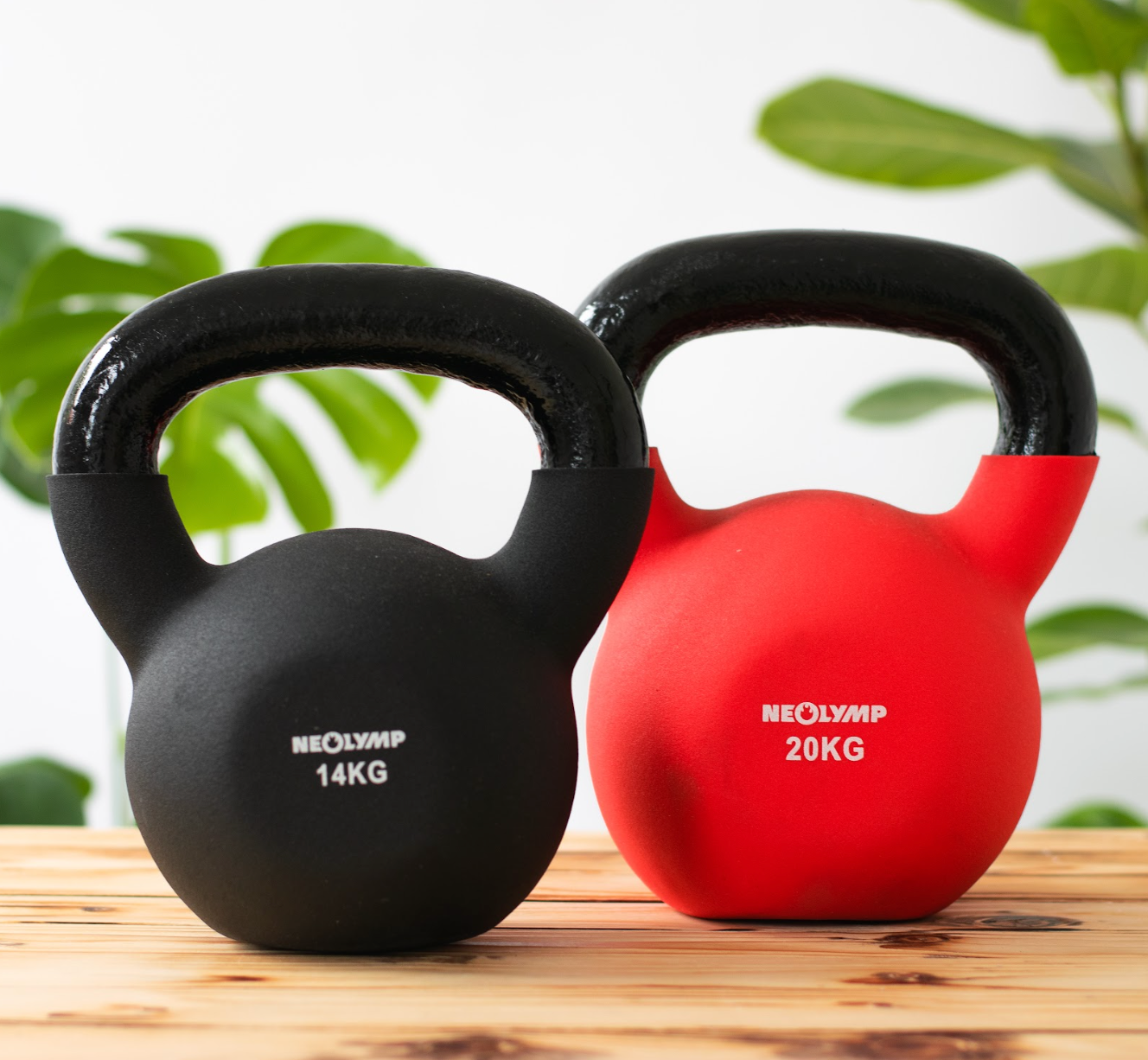 Zwei Neolymp Kettlebells stehen nebeneinander auf einer Holzoberfläche. Die linke Kettlebell ist schwarz und wiegt 14 kg, die rechte ist rot und wiegt 20 kg. Im Hintergrund sind grüne Pflanzen unscharf zu sehen, die einen frischen und natürlichen Look vermitteln.