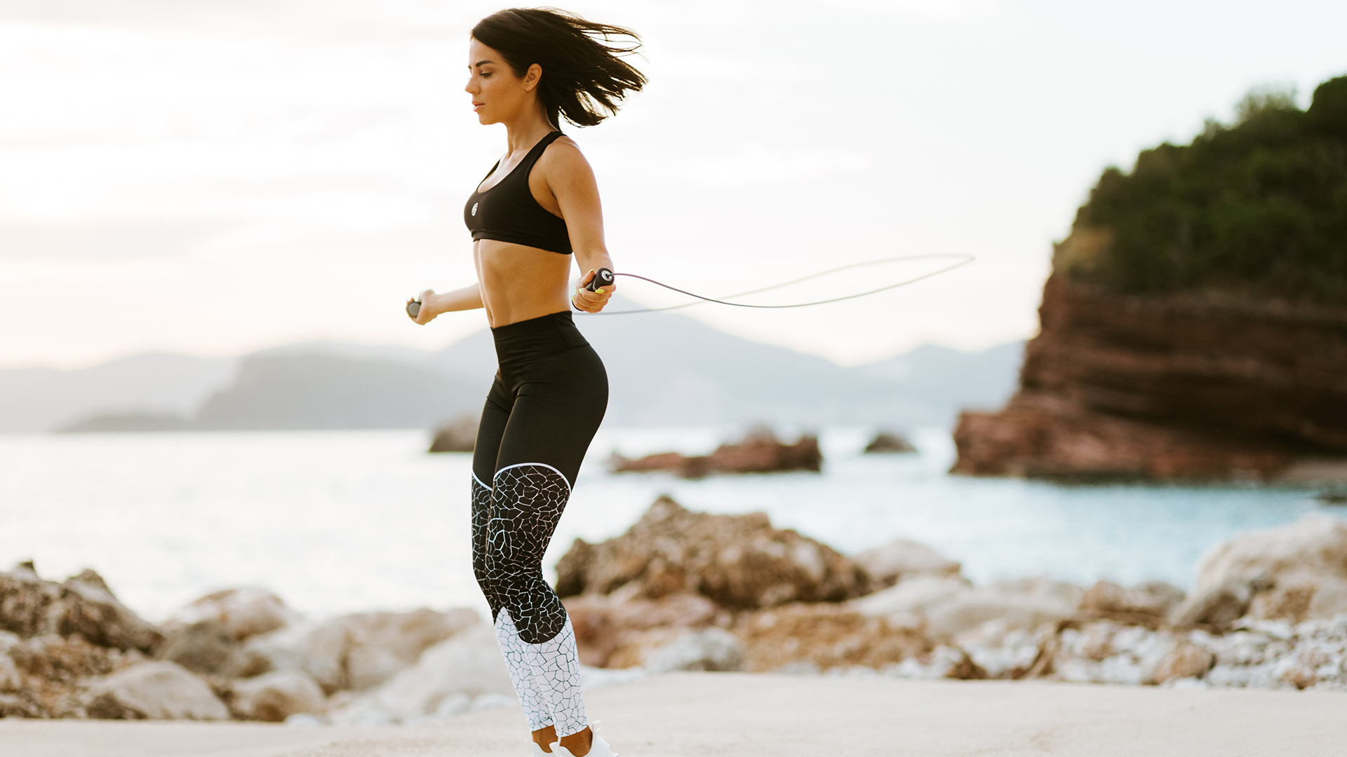 Frau trainiert mit einem Springseil am Strand bei Sonnenuntergang, fokussiert auf ihr Cardio-Workout vor einer malerischen Meereskulisse.