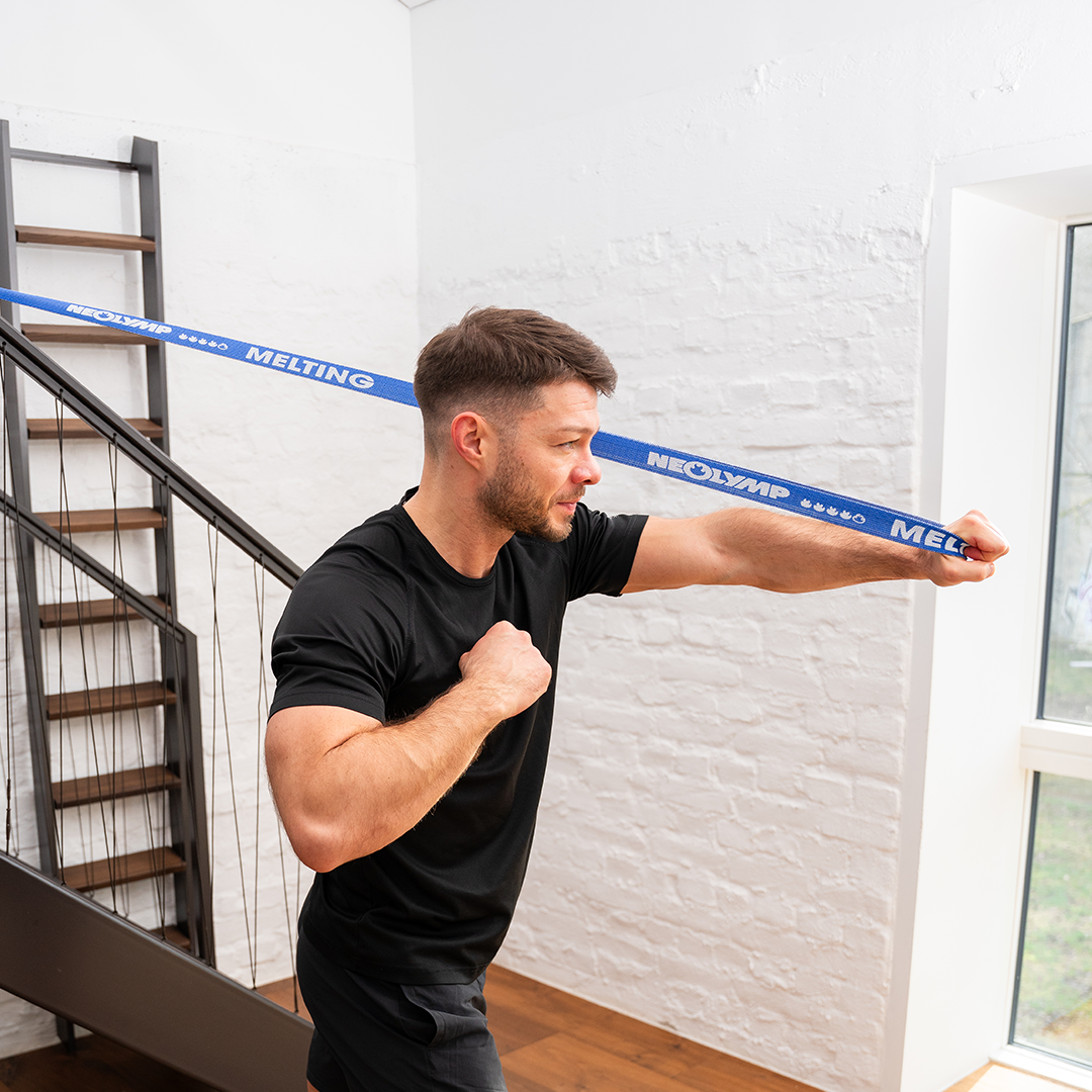 Ein Mann führt eine Widerstandsübung mit einem blauen Neolymp-Band (Melting) durch, das an einem Treppengeländer befestigt ist. Er trägt ein schwarzes T-Shirt und schwarze Shorts. Im Hintergrund ist eine weiße Ziegelwand und eine moderne Treppe zu sehen.